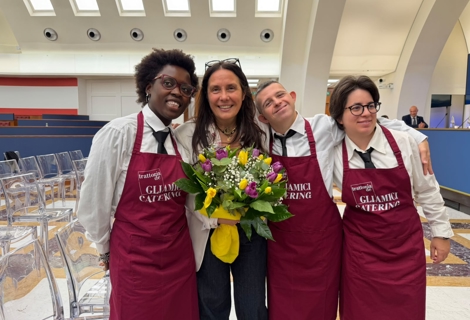 Il Ministro per le Disabilità Alessandra Locatelli posa insieme ad alcuni operatori della Trattoria degli Amici all’interno della sala della Presidenza del Consiglio dei Ministri. Le persone indossano i grembiuli bordeaux del servizio catering della cooperativa e sono raccolte attorno al Ministro, che tiene in mano un mazzo di fiori. Sullo sfondo sono visibili le sedute della sala e i lucernari della caratteristica copertura dell’aula